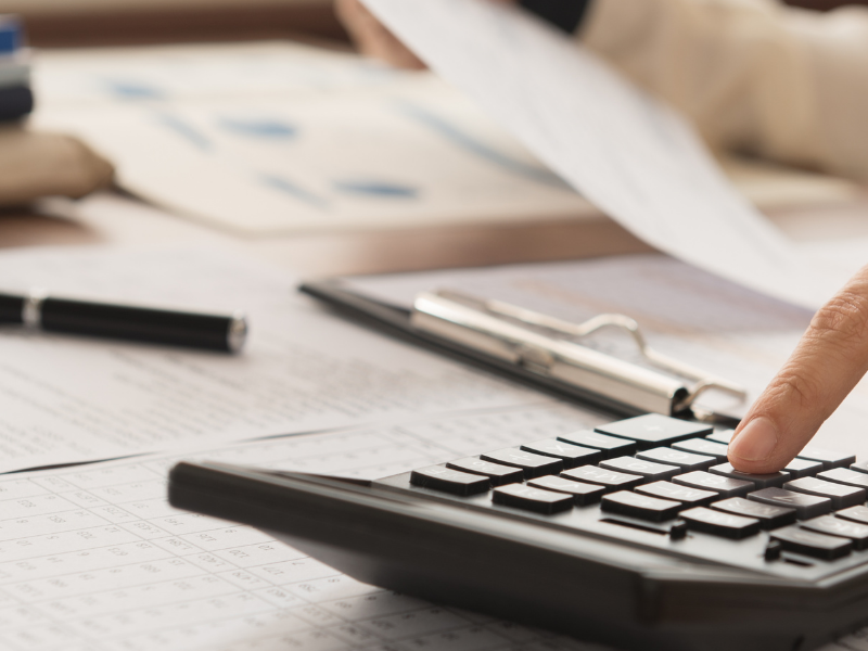 Person calculates finances with a calculator on a desk cluttered with papers, a clipboard, and a pen.
