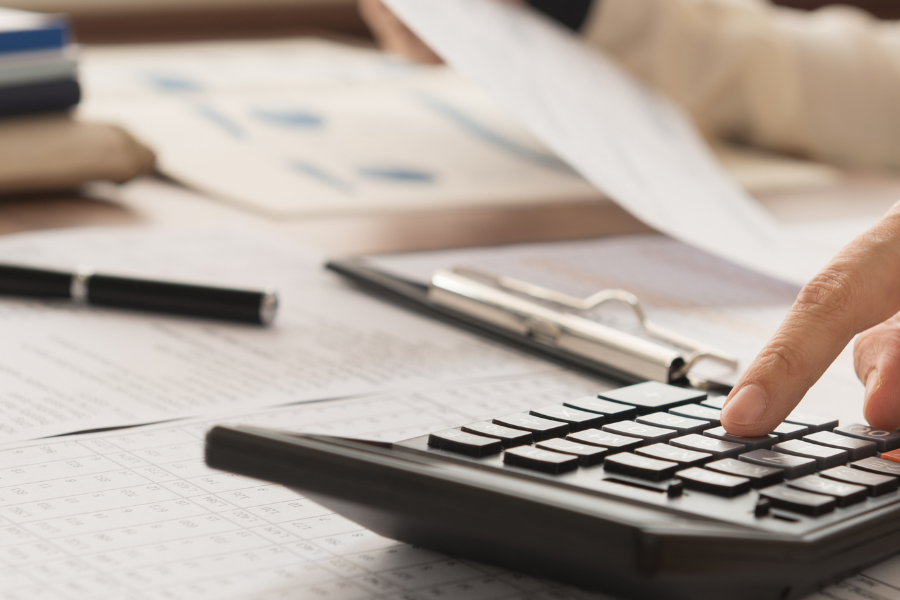 Person calculates finances with a calculator on a desk cluttered with papers, a clipboard, and a pen.