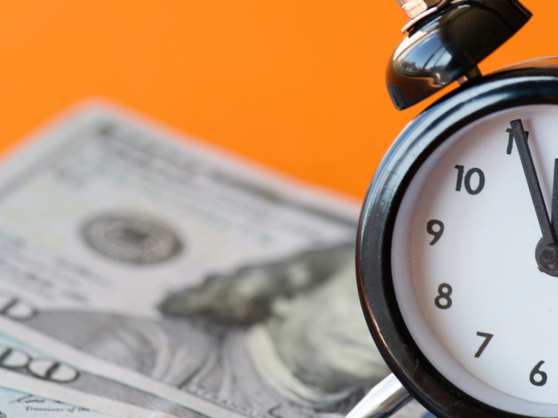 Analog alarm clock beside scattered $100 bills on an orange background, conveying the idea 'time is money' or financial urgency.