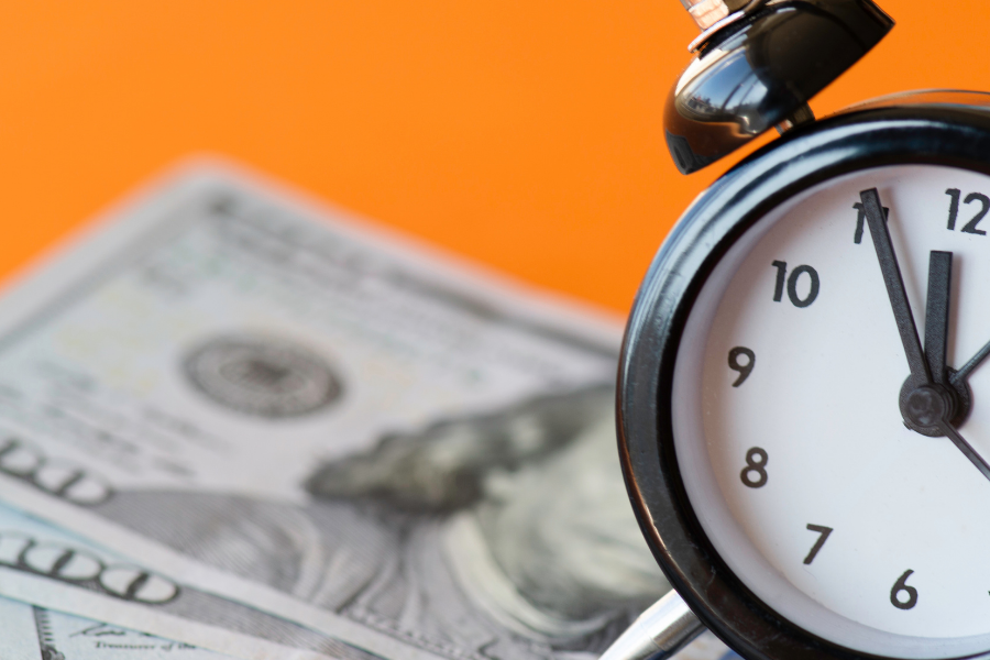 Analog alarm clock beside scattered $100 bills on an orange background, conveying the idea 'time is money' or financial urgency.