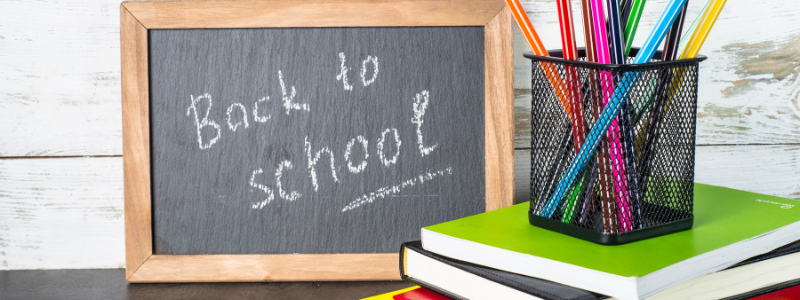 Chalkboard reading 'Back to school' beside a pencil holder and stack of notebooks on a wooden desk.