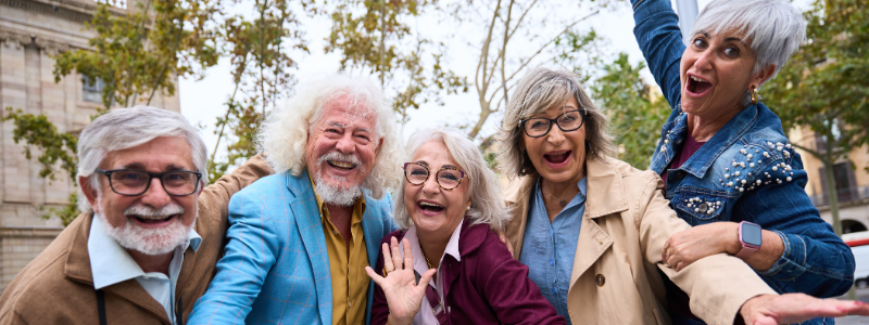 Five smiling seniors posing together outdoors, arms around each other and waving at the camera.